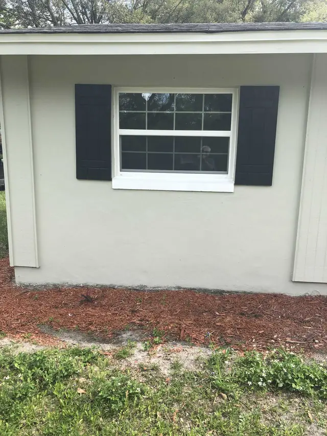 White-framed grid window between black shutters on a light exterior wall.